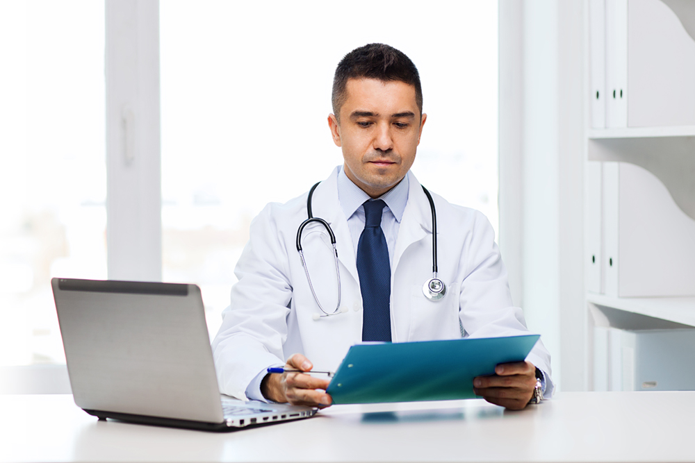 Male doctor with clipboard and laptop computer in medical office